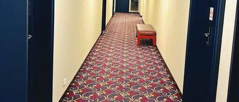 A hotel hallway with a patterned carpet, doors on both sides, and a bench in the center.