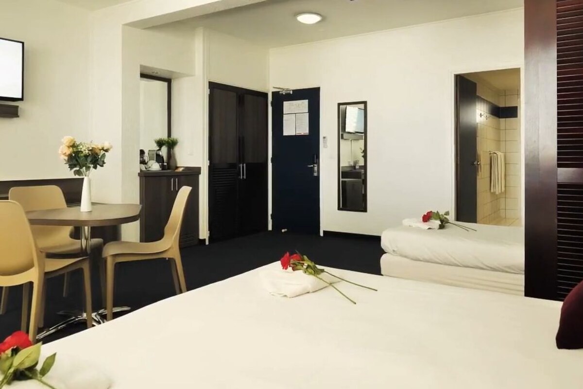 A hotel room with a bed, a desk, and a chair. There is a clock on the wall and a TV mounted above the desk.
