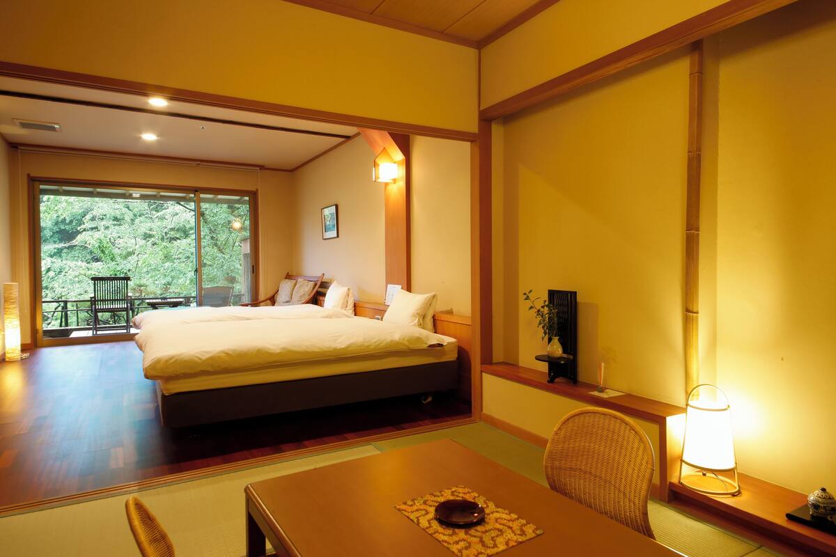 A hotel room with a large bed, a dining table, a chair, and a view of greenery outside.