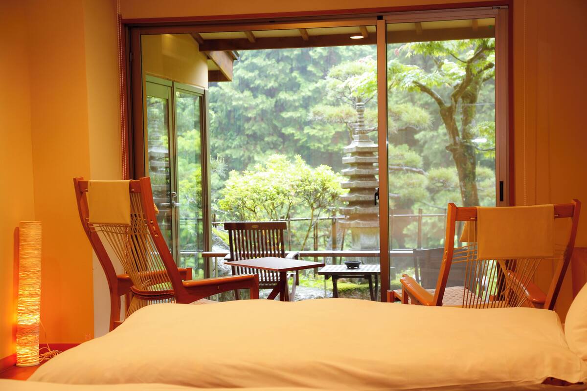 A room with a bed, two wooden chairs, a small table, and a view of a pagoda and trees through a large sliding glass door.