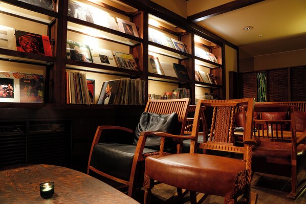 Una biblioteca con estanterías de madera llenas de libros y discos de vinilo. Hay varias sillas de madera y una mesa con una vela.