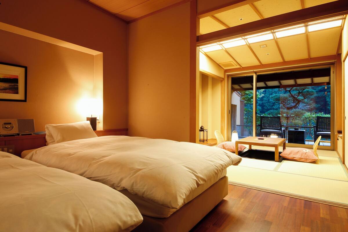A hotel room with a large bed, a small table, and a view of a garden through a sliding door.