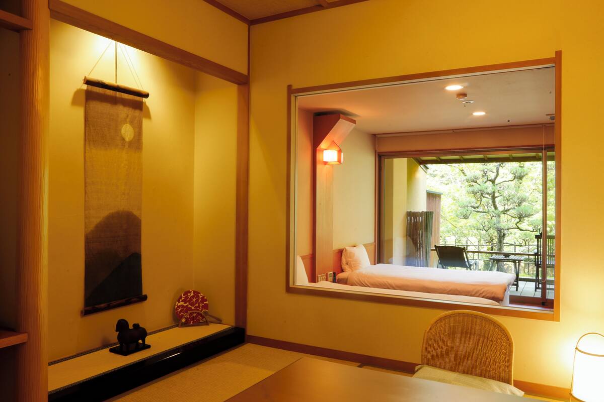 A hotel room with a bed, a chair, a small table, a wall-mounted shelf with decorative items, and a window with a view of the outdoors.