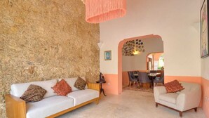 A living room with a stone wall, a white sofa with patterned cushions, a small table, and a dining area with chairs. - CASA DE LOS PLATOS, IN THE HEART OF MÉRIDA (Centro)