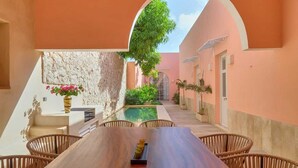 A wooden dining table with wicker chairs, a stone wall, a pool, and a view of a garden through an archway. - CASA DE LOS PLATOS, IN THE HEART OF MÉRIDA (Centro)