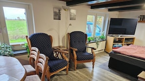Un salon avec un plancher en bois, deux fauteuils en bois recouverts d’un tissu bleu, une table ronde en bois, un téléviseur et une bibliothèque.