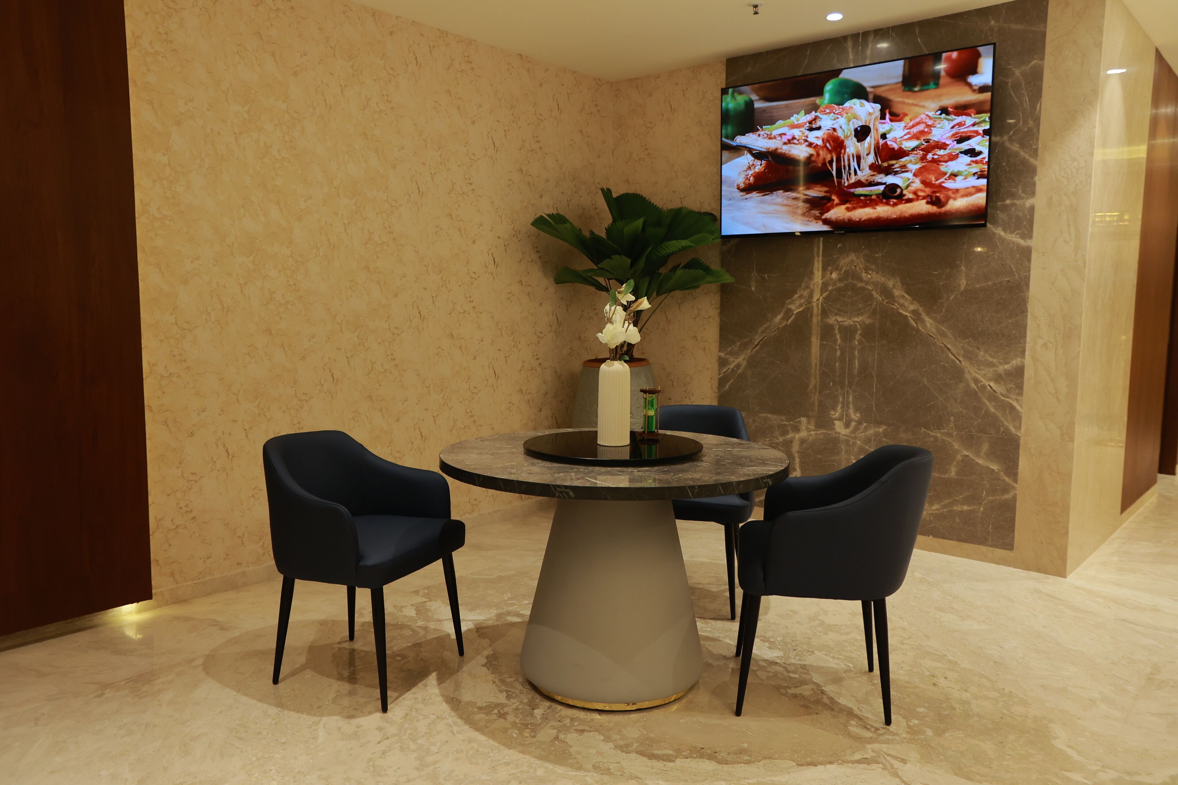 Un comedor moderno con una mesa de mármol, cuatro sillas oscuras, una planta en maceta y un televisor de pantalla plana montado en la pared.