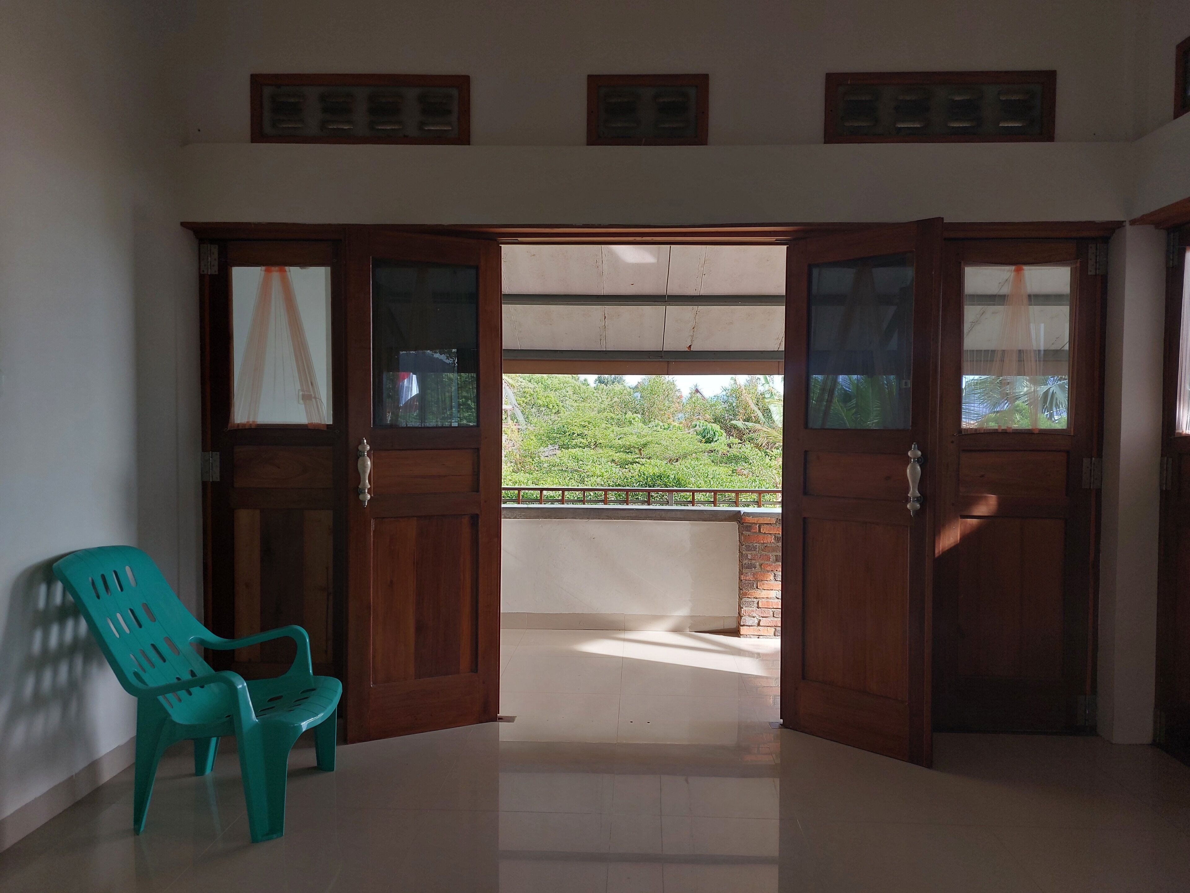 Een kamer met een houten deur die leidt naar een balkon, een turquoise plastic stoel en een betegelde vloer.