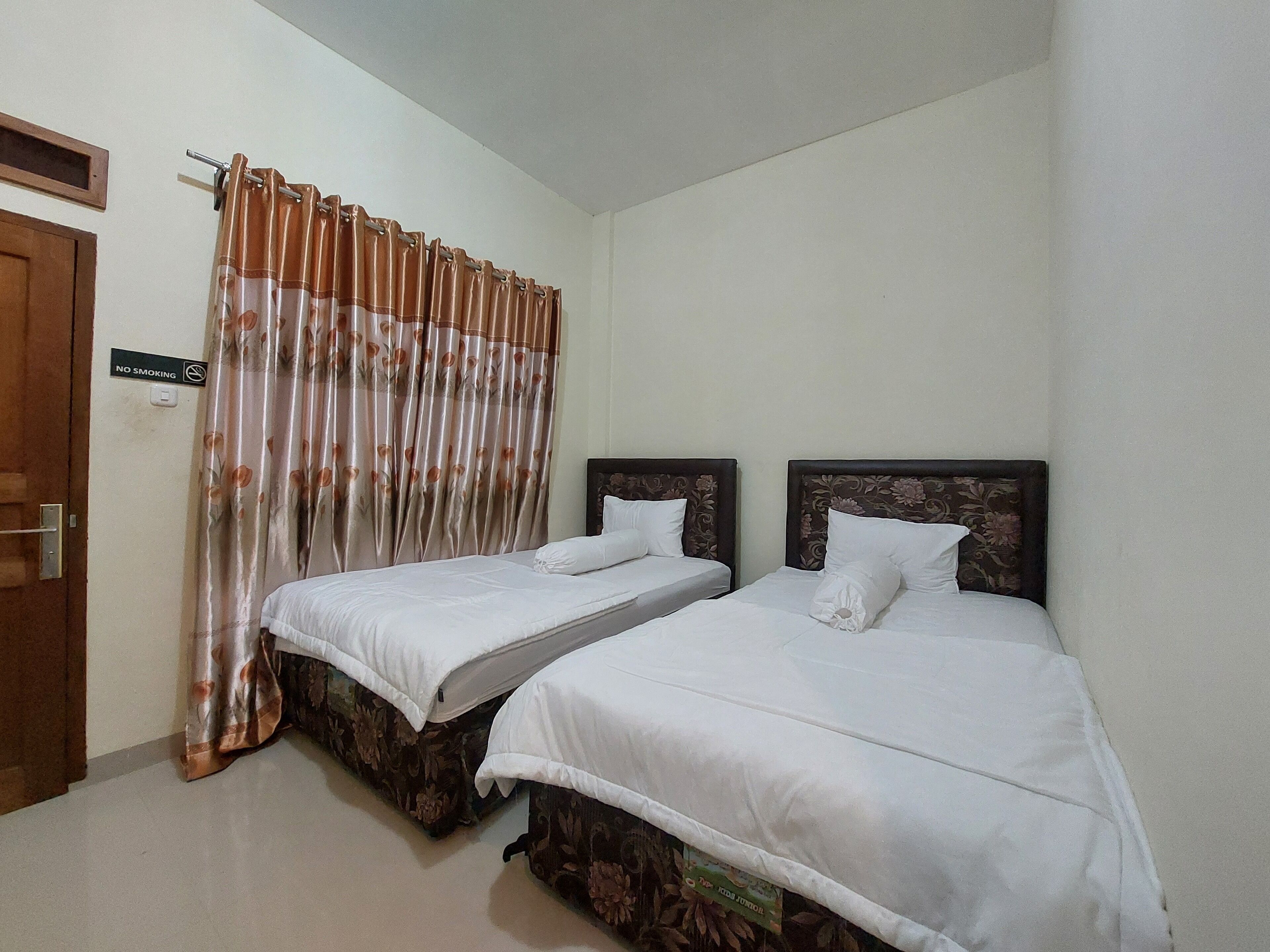 A room with two single beds, a wooden door, a 'No Smoking' sign, and a curtain with a floral pattern.