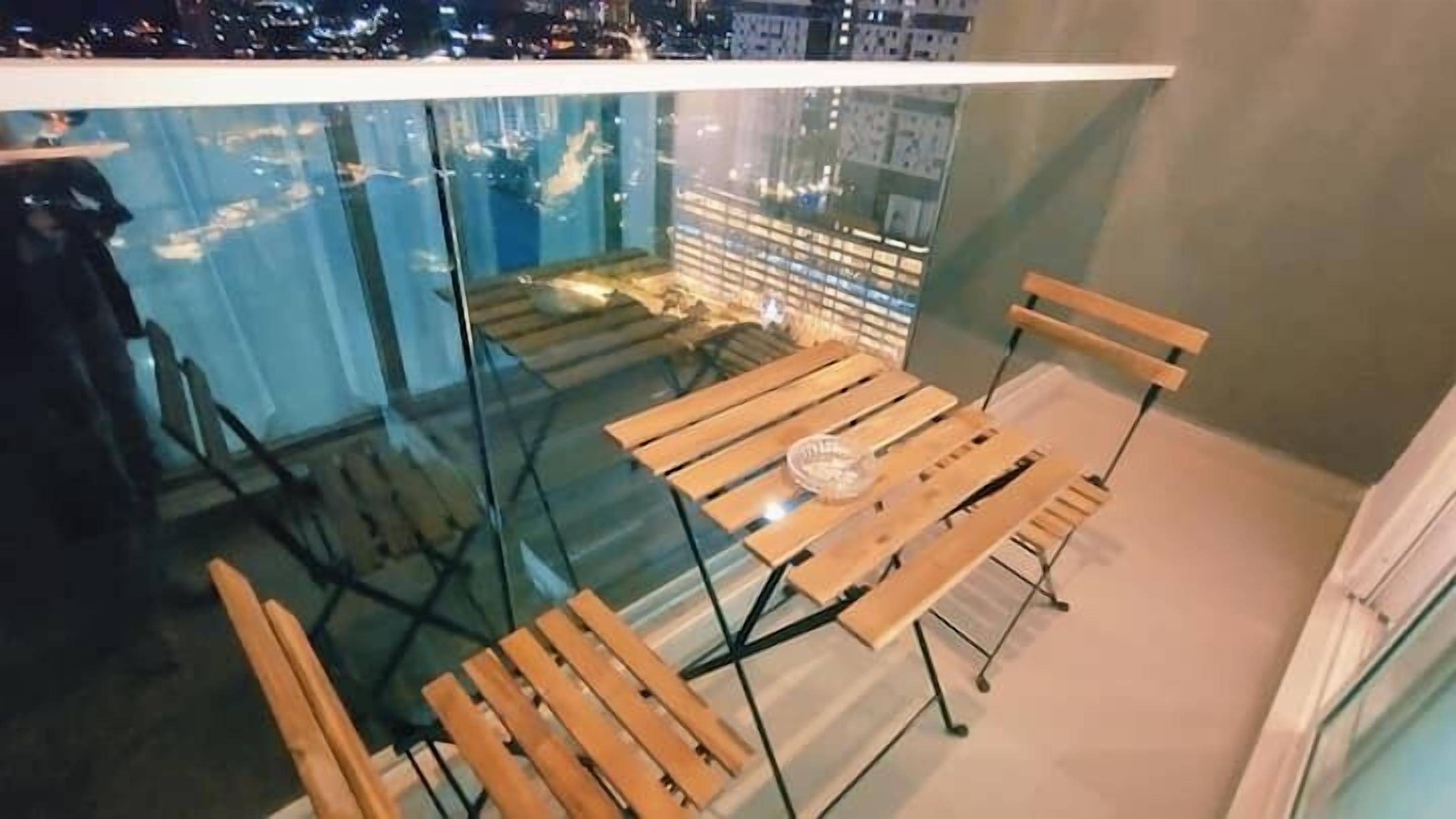 Un balcón con baranda de vidrio, sillas de madera y una mesa.