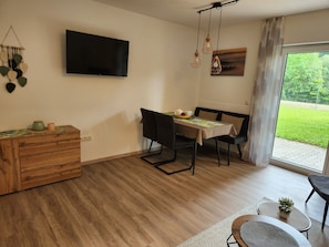 A modern living room with a flat-screen TV, a dining table with chairs, a wooden cabinet, and a view of a green landscape through a large window. - Holiday Apartment '5 Traminer' with Mountain View, Shared Terrace and Wi-Fi (Iphofen)