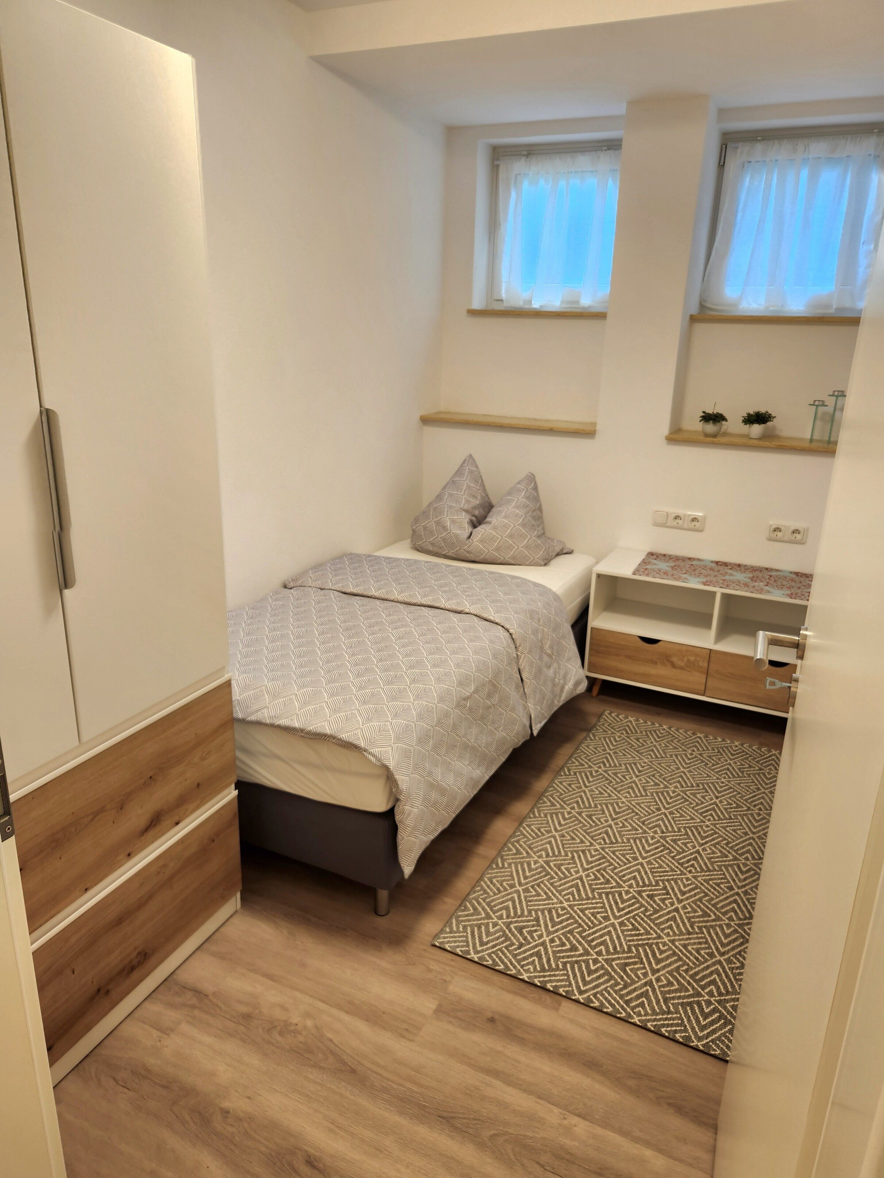 A small, neatly arranged bedroom with a bed, bedside table, and a window.