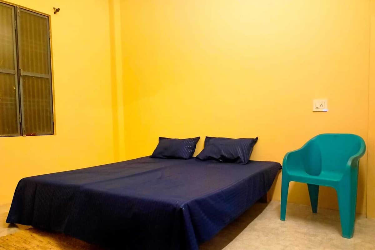 Una cama individual con una colcha azul oscuro y dos almohadas, una silla de plástico color turquesa y una ventana con marco de metal.