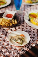 Una mesa con mantel estampado, un tazón de sopa con verduras, un plato de papas fritas y una bebida azul.