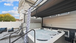 Un jacuzzi con baranda metálica, un patio cubierto con mobiliario exterior, y una casa de fachada gris.
