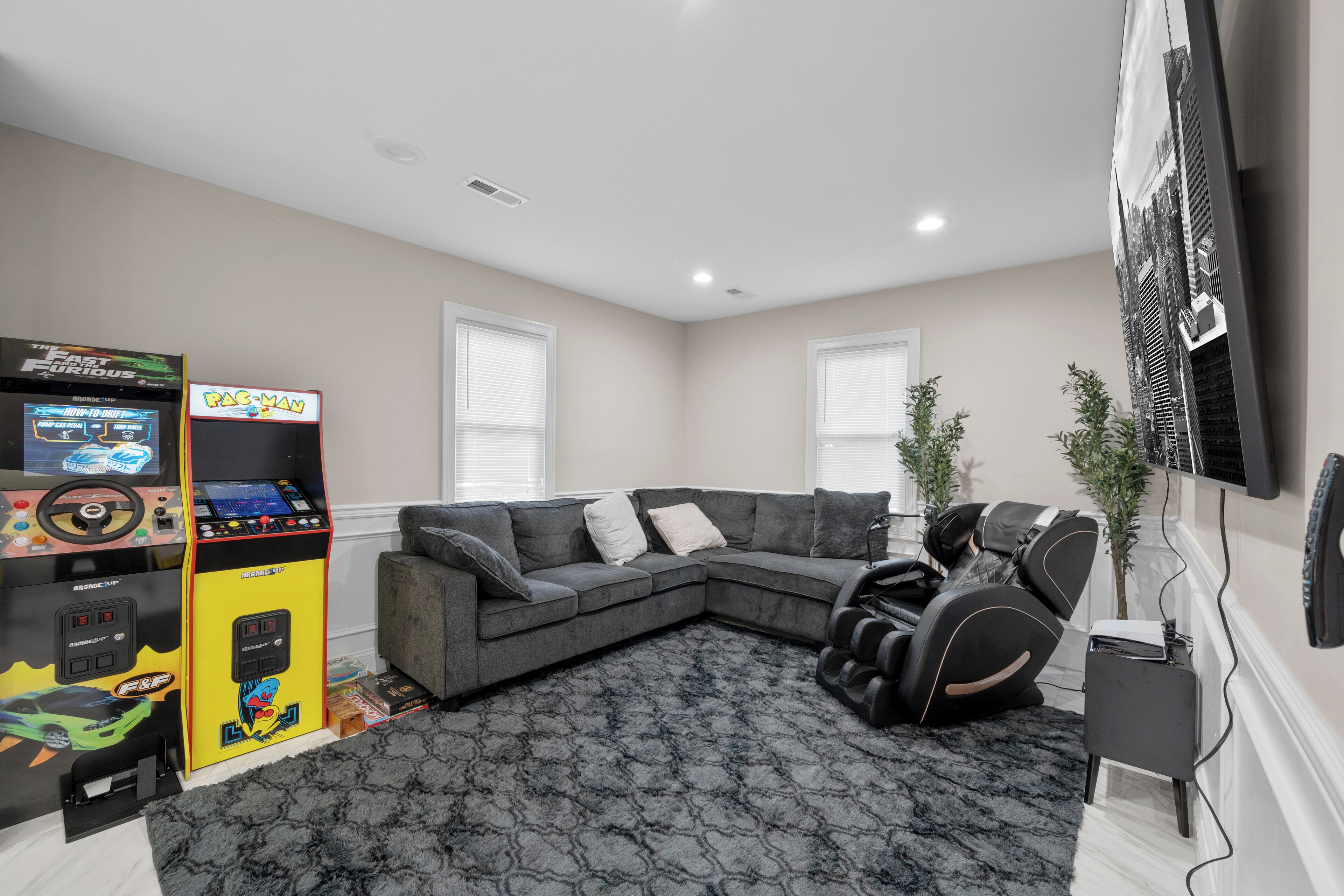 A living room with a gray sectional sofa, a massage chair, a large flat-screen TV mounted on the wall, and a Pac-Man arcade machine.