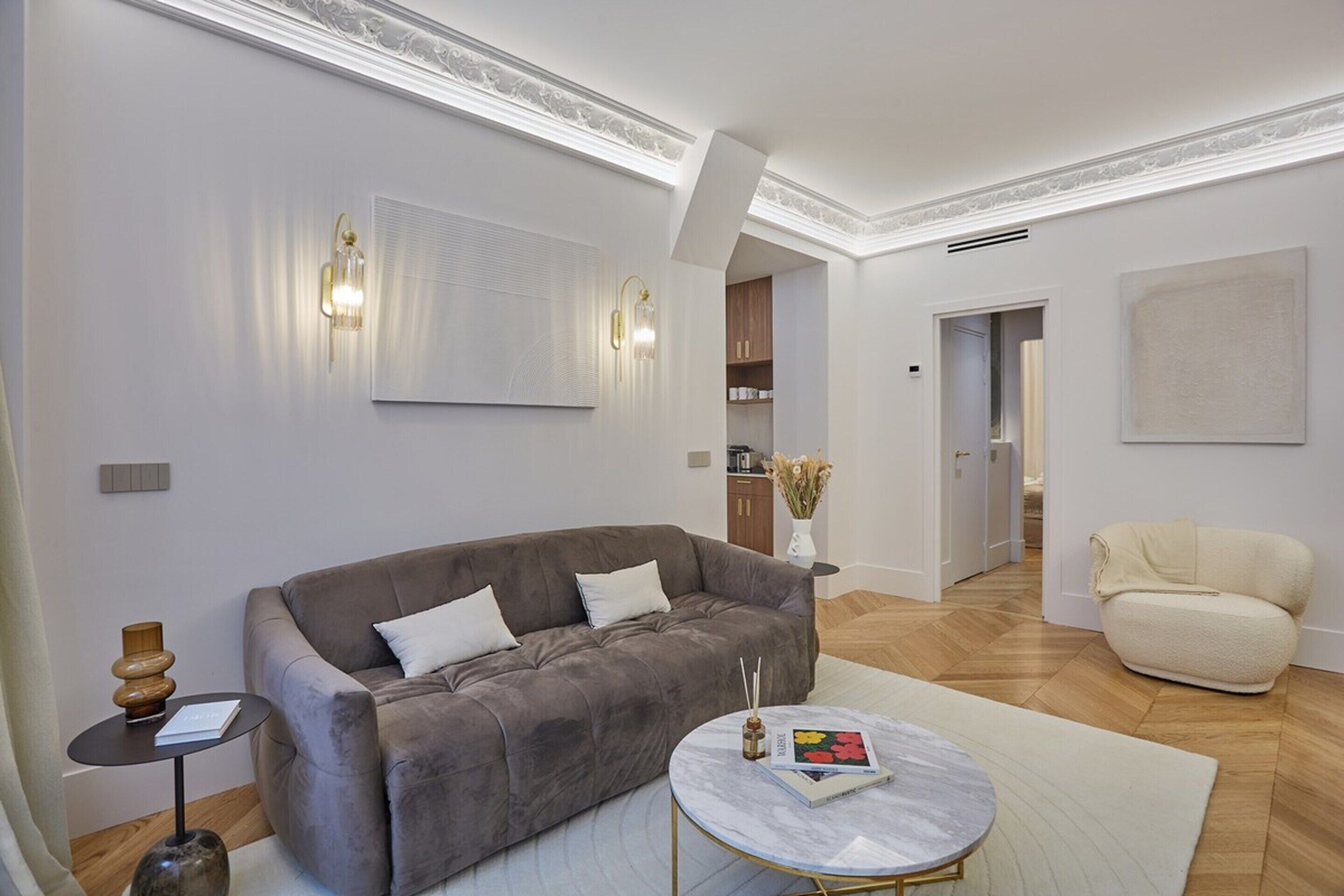 A modern living room with a grey sofa, a round coffee table, and a beige armchair. There is a bookshelf with books and decorative items, and a large abstract painting on the wall.