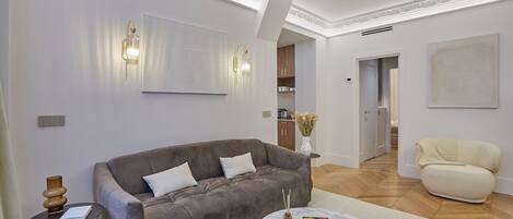 A modern living room with a grey sofa, a round coffee table, and a beige armchair. There is a bookshelf with books and decorative items, and a large abstract painting on the wall.