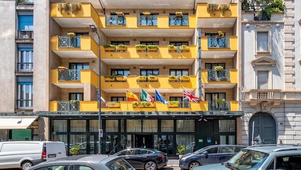 A multi-story hotel with balconies, flags, and a parking lot. - Mythos (Milan)