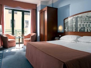 A hotel room with a large bed, a desk with a chair, a wardrobe, and a balcony with a view of buildings. - Mythos (Milan)