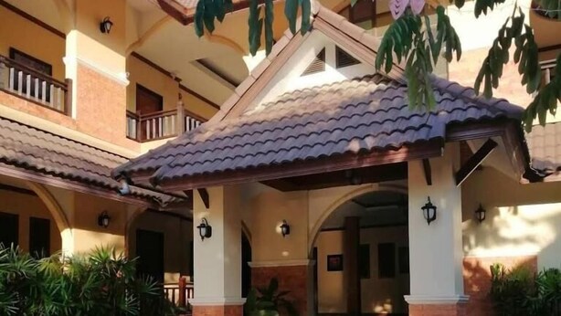 Uma área de estar coberta ao ar livre com móveis de vime e uma pequena mesa, que dá acesso a um portal de edifício com telhado em azulejos e colunas.
