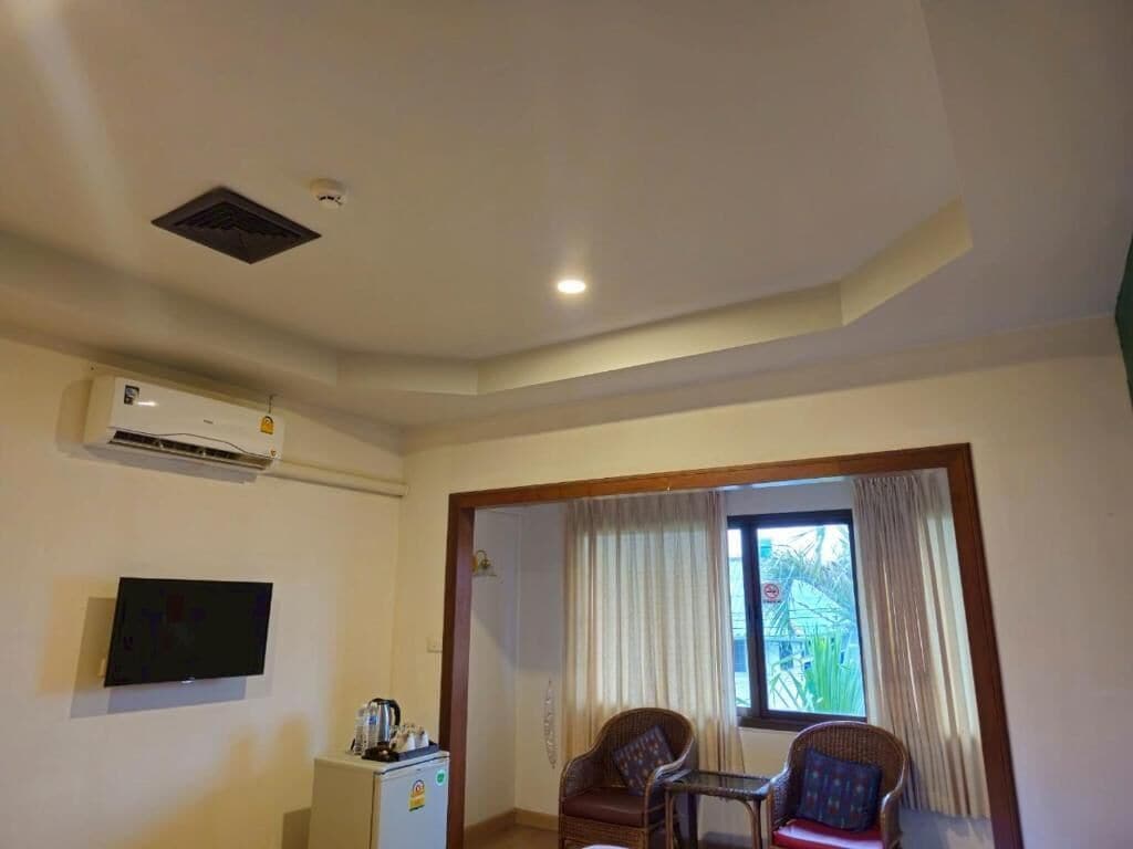 Una habitación con televisión de pantalla plana en la pared, una pequeña mesa con sillas, una ventana con cortinas y un equipo de aire acondicionado.