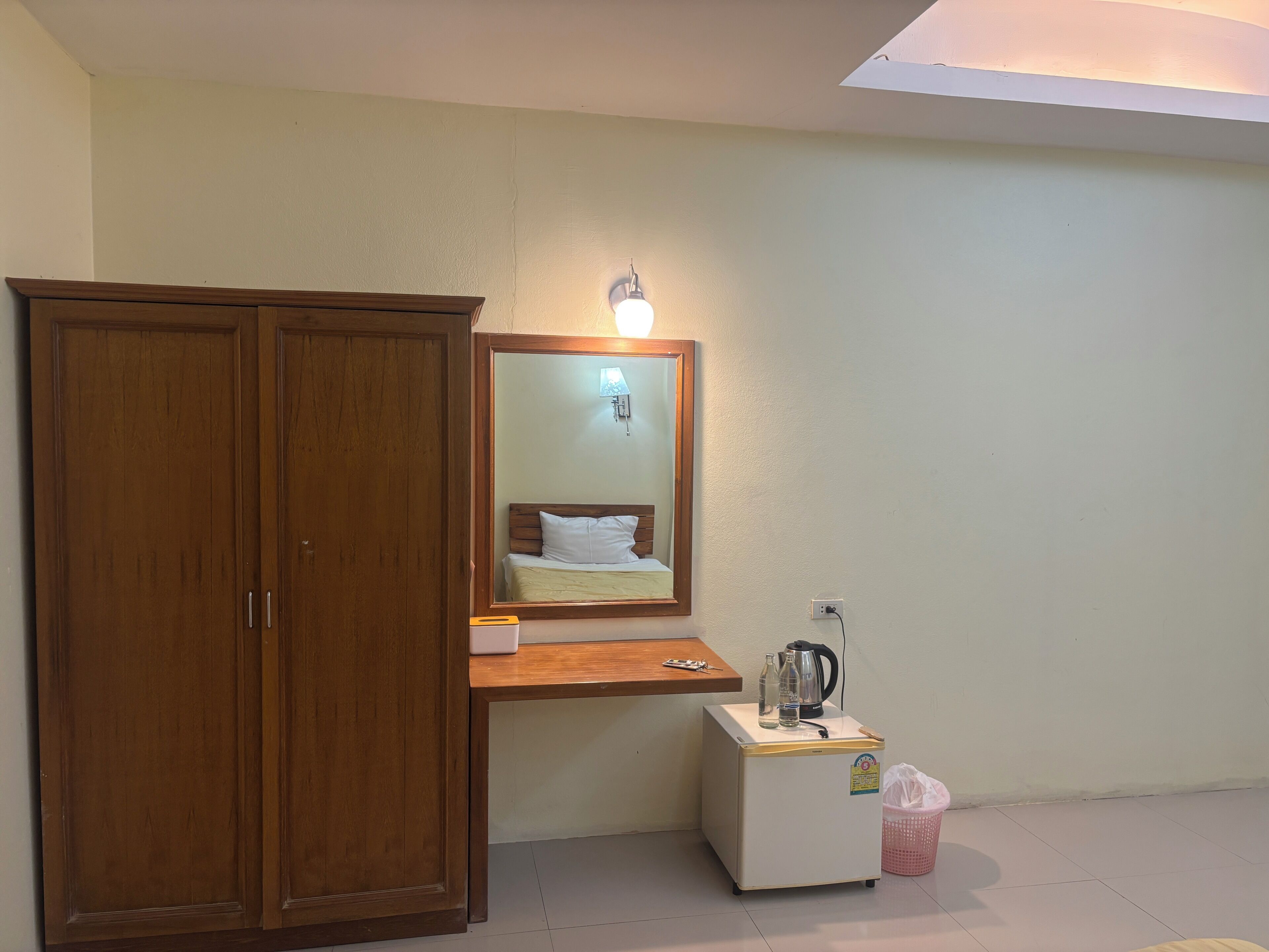 A room with a wooden wardrobe, a small white refrigerator, a mirror with a light, and a wall socket.