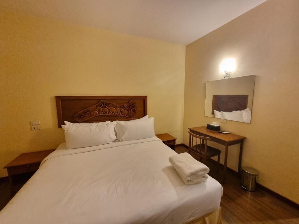 A hotel room with a large bed, a wooden headboard, a bedside table, a desk, a chair, and a mirror on the wall.
