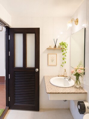 Un baño con una puerta de madera oscura, un lavabo blanco con encimera de mármol, un espejo, un cuadro enmarcado, una planta en maceta y un dispensador de papel higiénico.