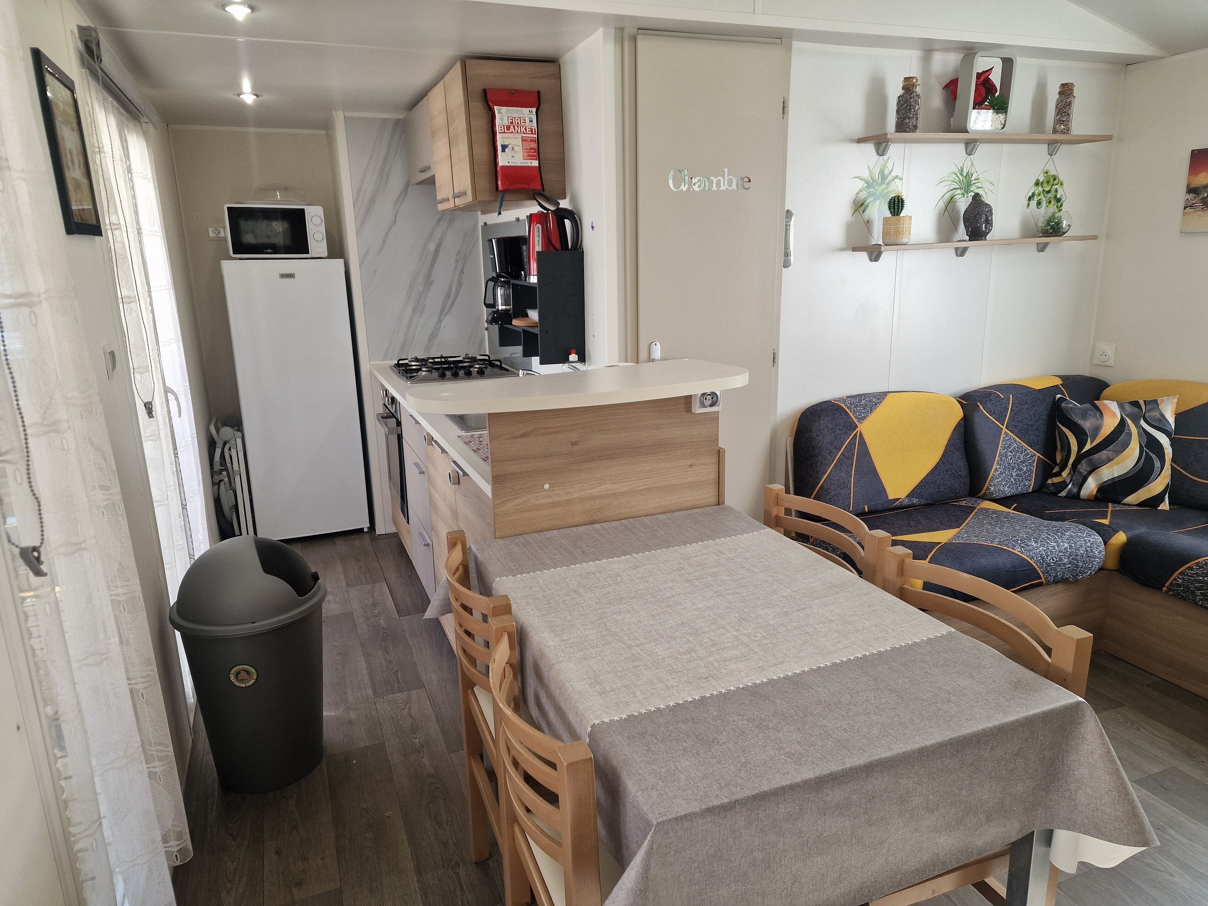 A compact kitchen with a microwave, stove, and coffee maker, a dining table with chairs, and a sofa with patterned cushions.