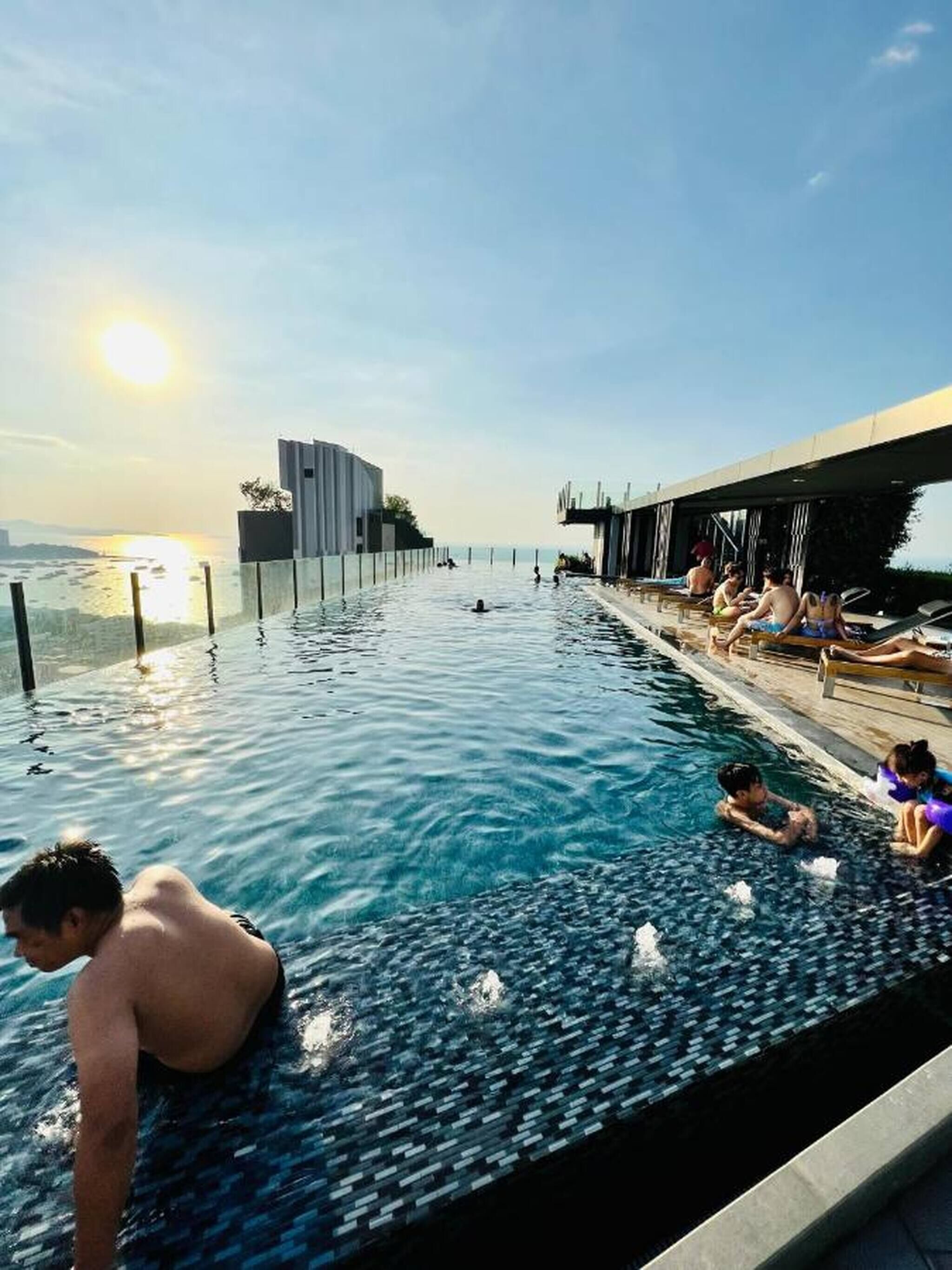 An infinity pool with a view of the sunset, a modern building, and lounge chairs along the edge.