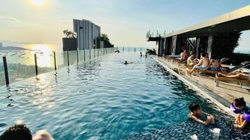 An infinity pool with a view of the sunset, a modern building, and lounge chairs along the edge.