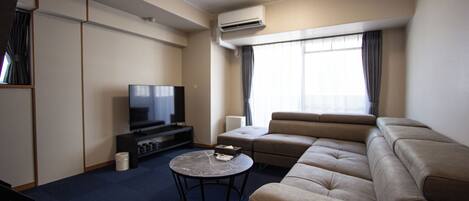 A modern hotel room with a large sofa, a flat-screen TV, a coffee table, and a large window with curtains.