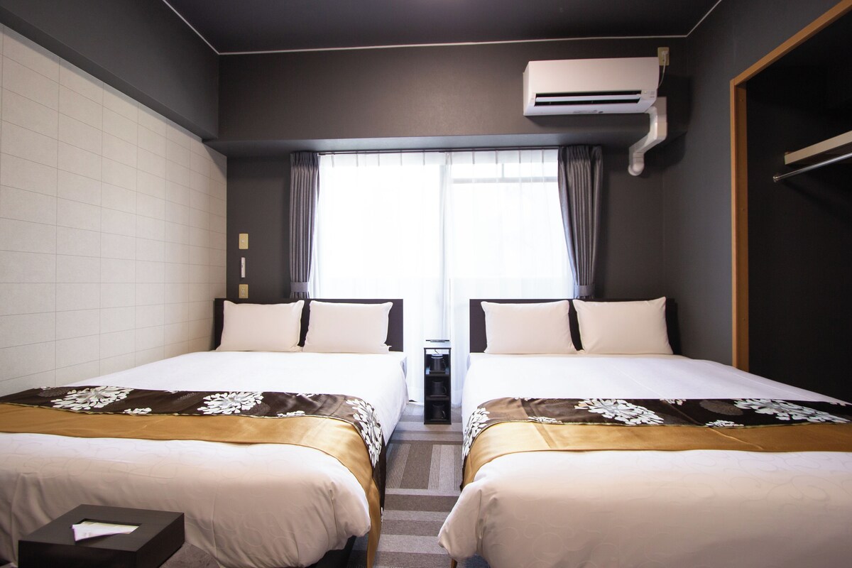 A hotel room with two beds, a window with curtains, an air conditioning unit, and a small table with a box.