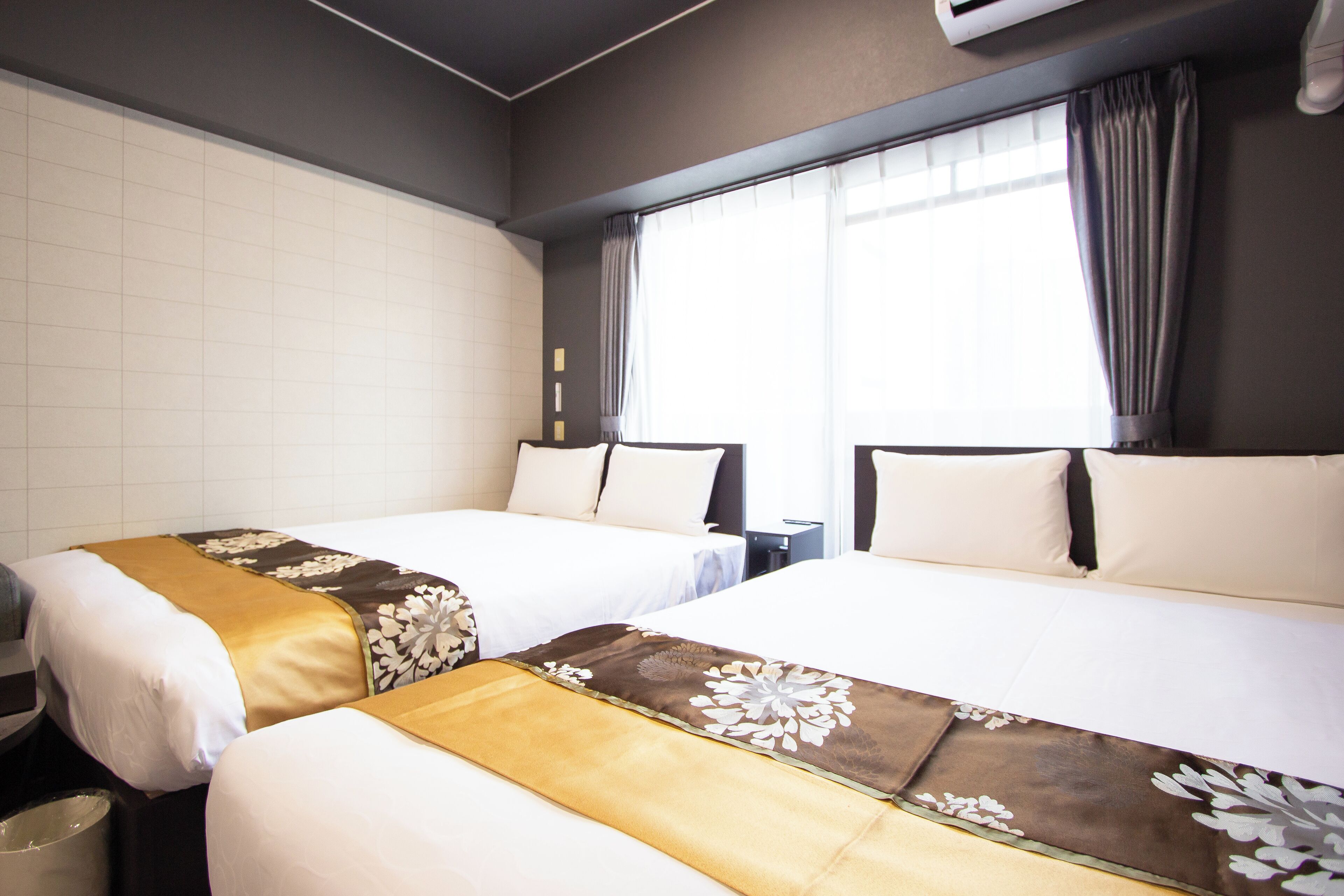 Een hotelkamer met twee bedden, elk voorzien van een bloemenprint deken in wit en bruin, een klein nachtkastje en een raam met gordijnen.