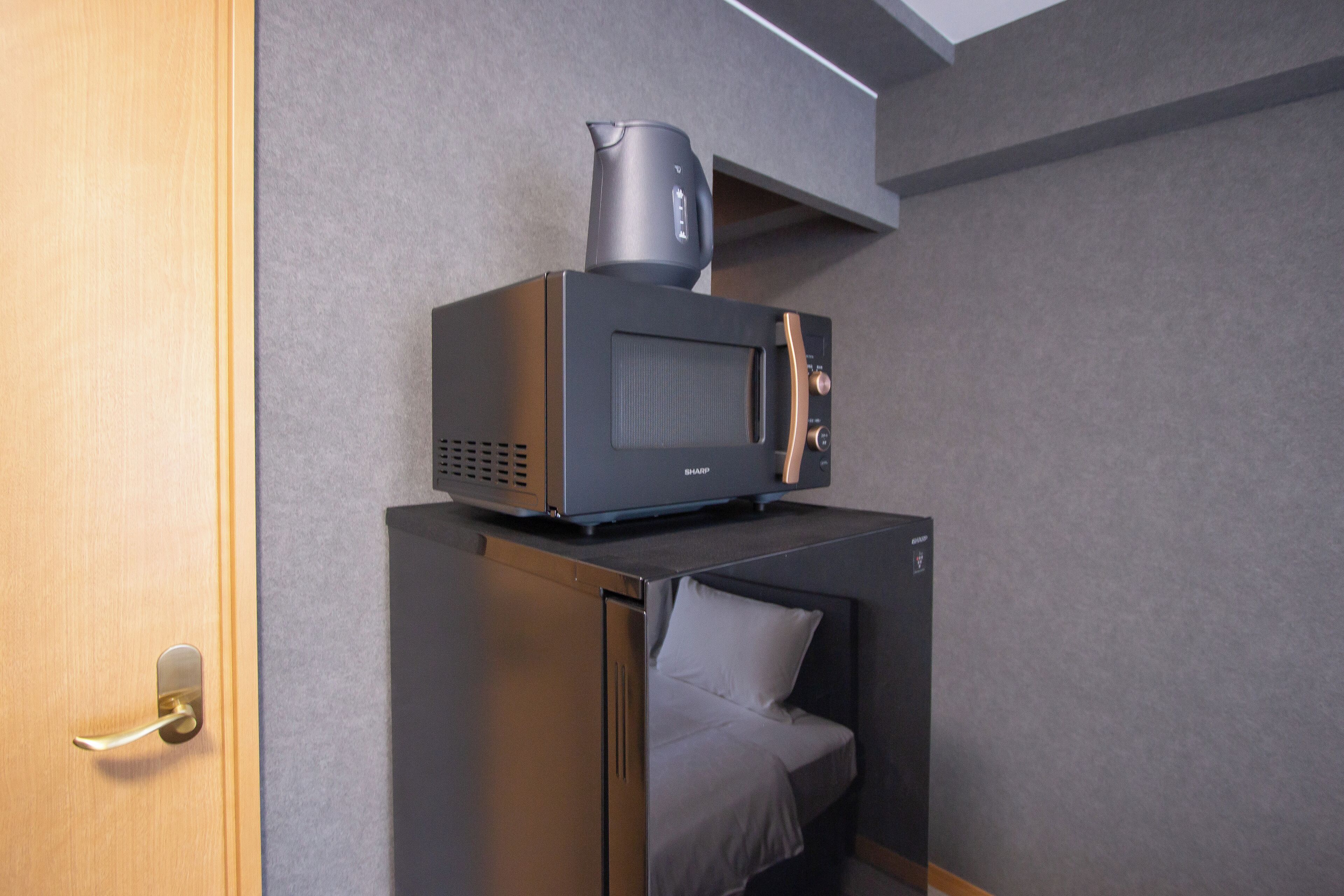A compact kitchenette with a microwave and coffee maker, a bed with a pillow, and a wooden door.