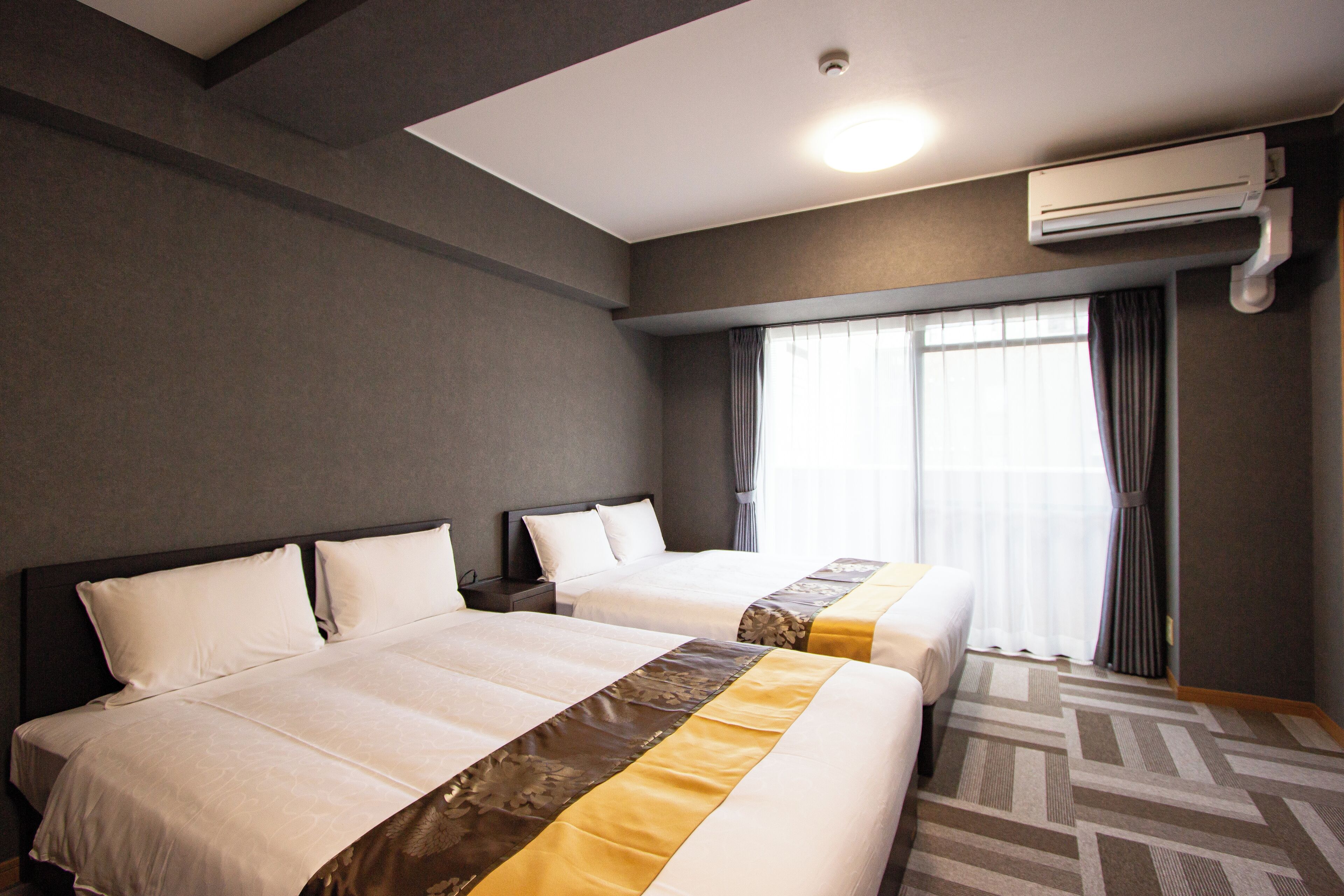 A hotel room with two beds, a large window with curtains, an air conditioning unit, and a patterned carpet.