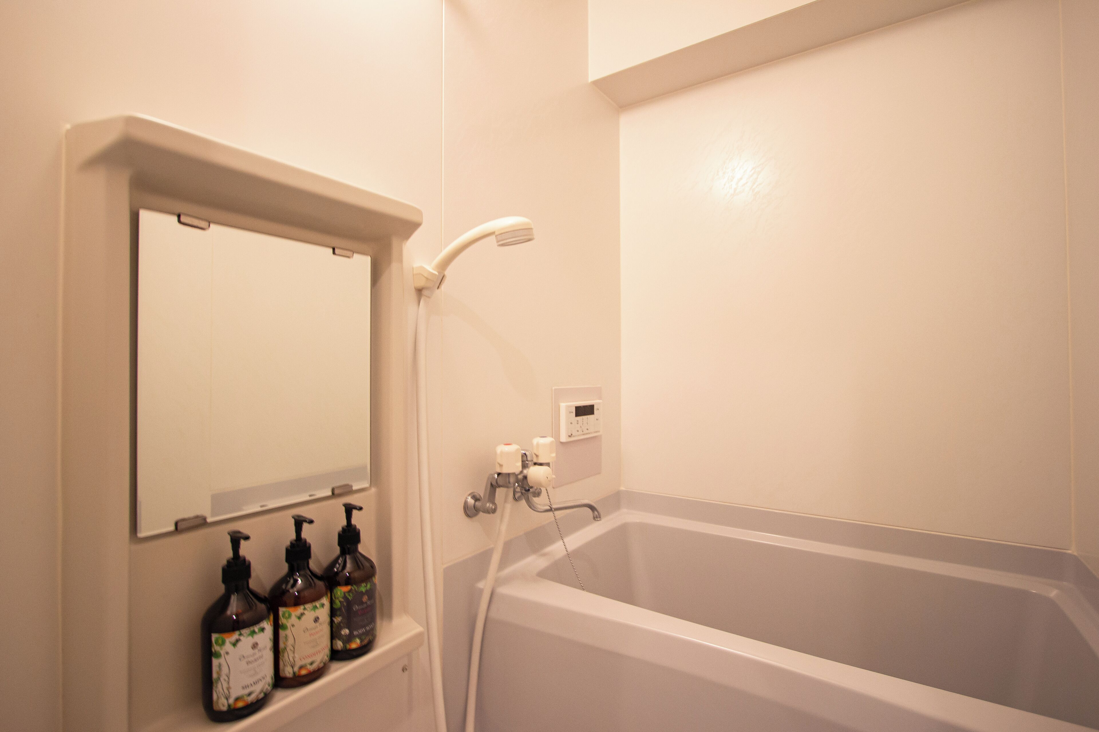 A bathroom with a bathtub, a wall-mounted mirror, and three bottles of liquid soap on a shelf.