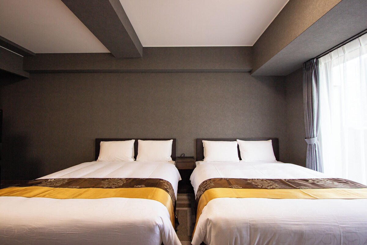 A hotel room with two single beds, a nightstand, a window with curtains, and a neutral color scheme.