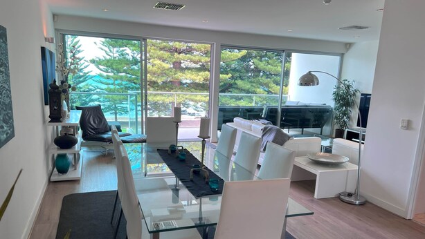 A modern living room with a glass dining table, white sofas, and a large window offering a view of trees and a car.
