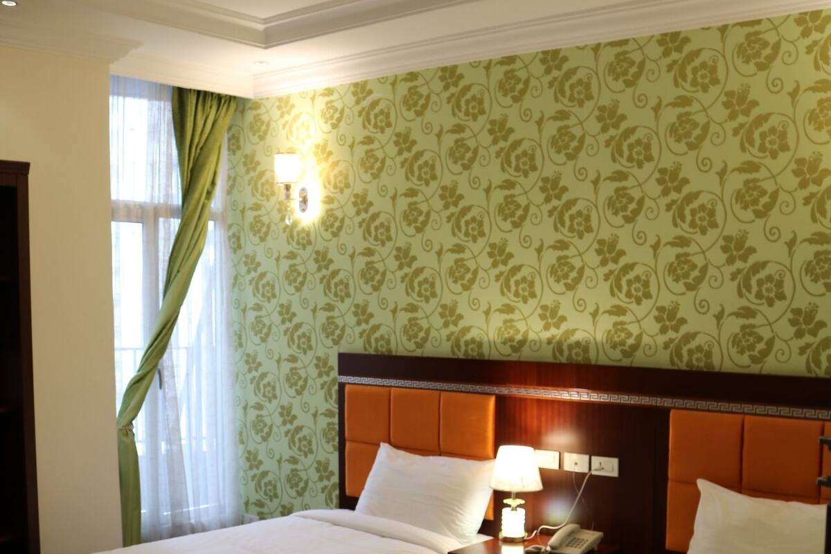 A hotel room with two beds, a nightstand with a lamp, a telephone, and a window with curtains.