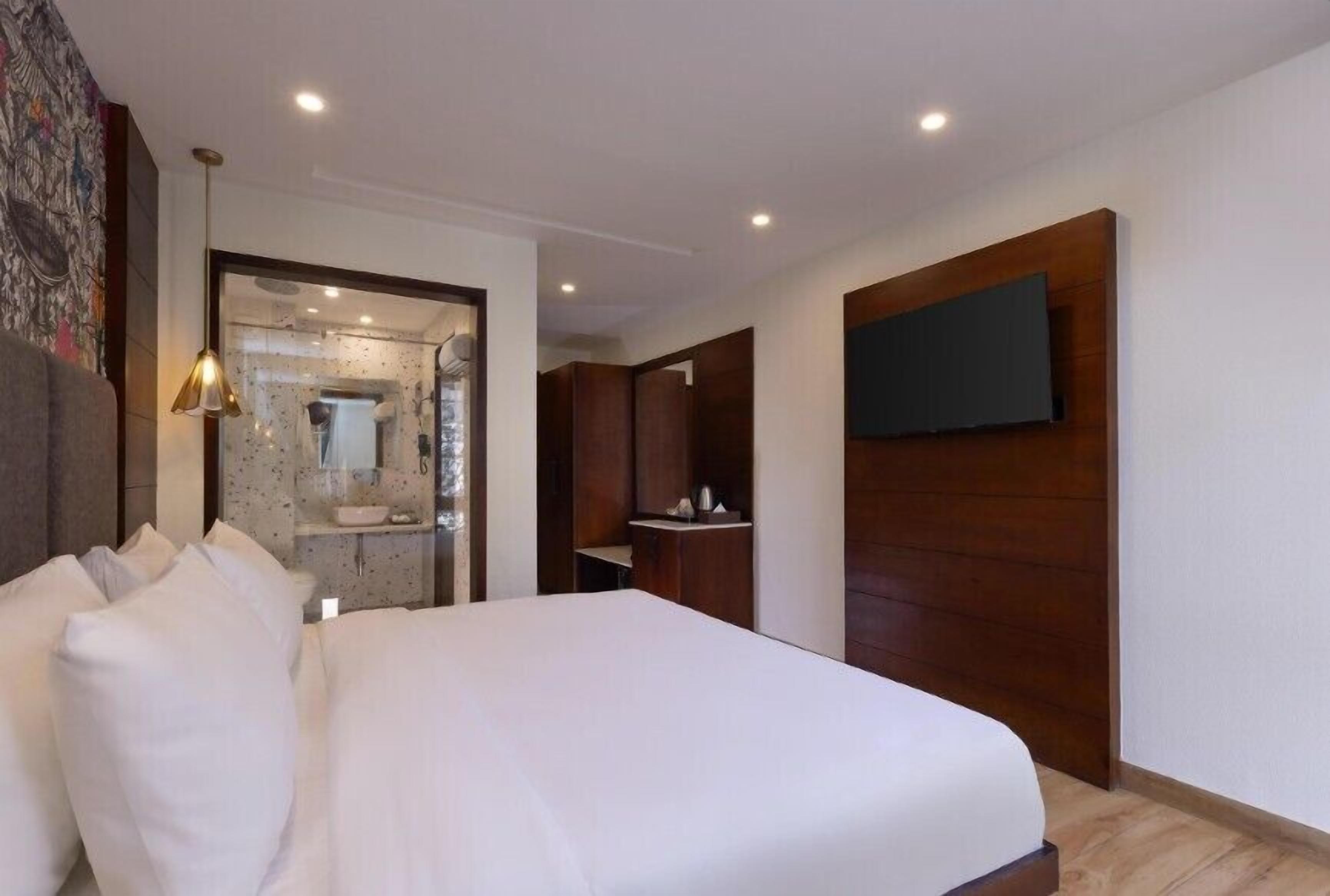 A hotel room with a large bed, a television mounted on the wall, a wooden wardrobe, and a bathroom visible through an open door.