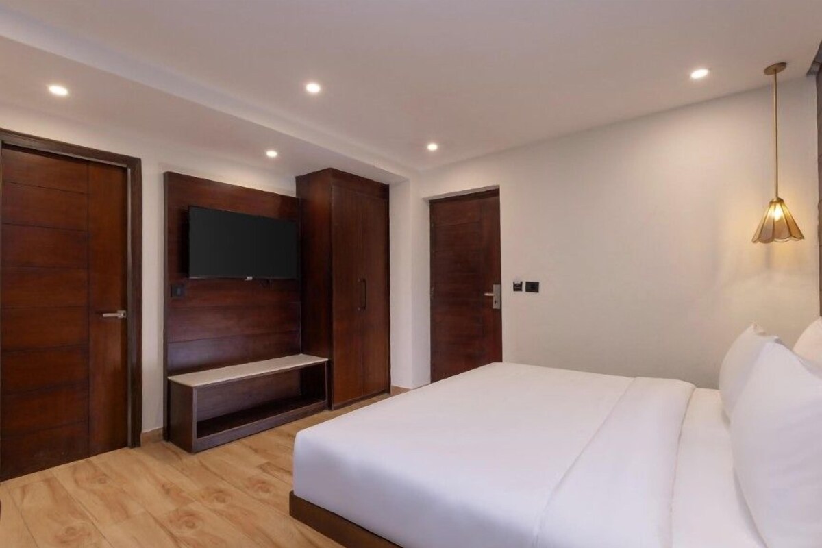 A bedroom with a bed, a TV mounted on a wooden panel, a wardrobe, and a pendant light.