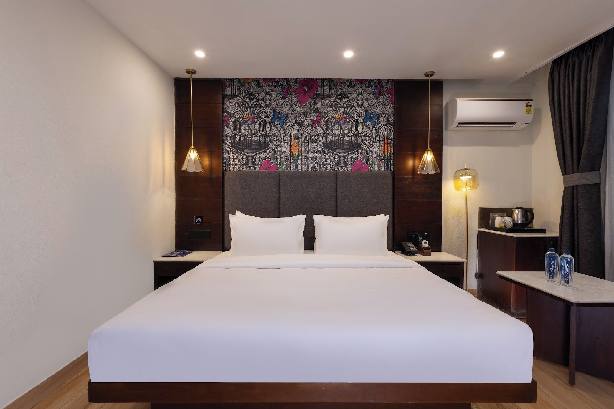A hotel room with a large bed, two bedside tables, a nightstand, an air conditioning unit, a lamp, and a decorative tapestry on the wall.