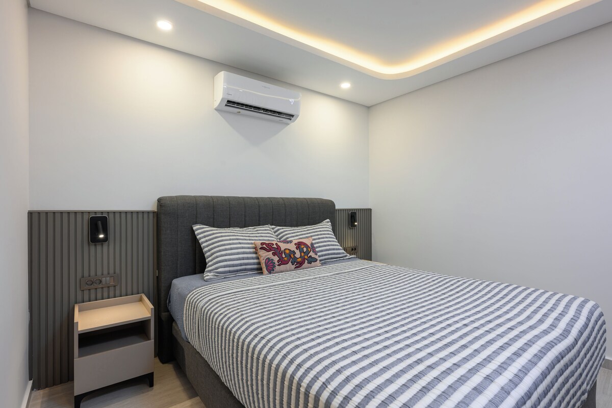 A modern bedroom with a bed, bedside tables, an air conditioning unit, and a wall-mounted light fixture.