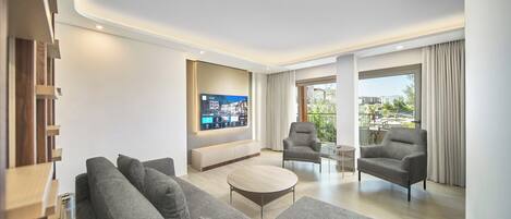A modern living room with a flat-screen TV, grey sofas, a coffee table, and a balcony with a view.
