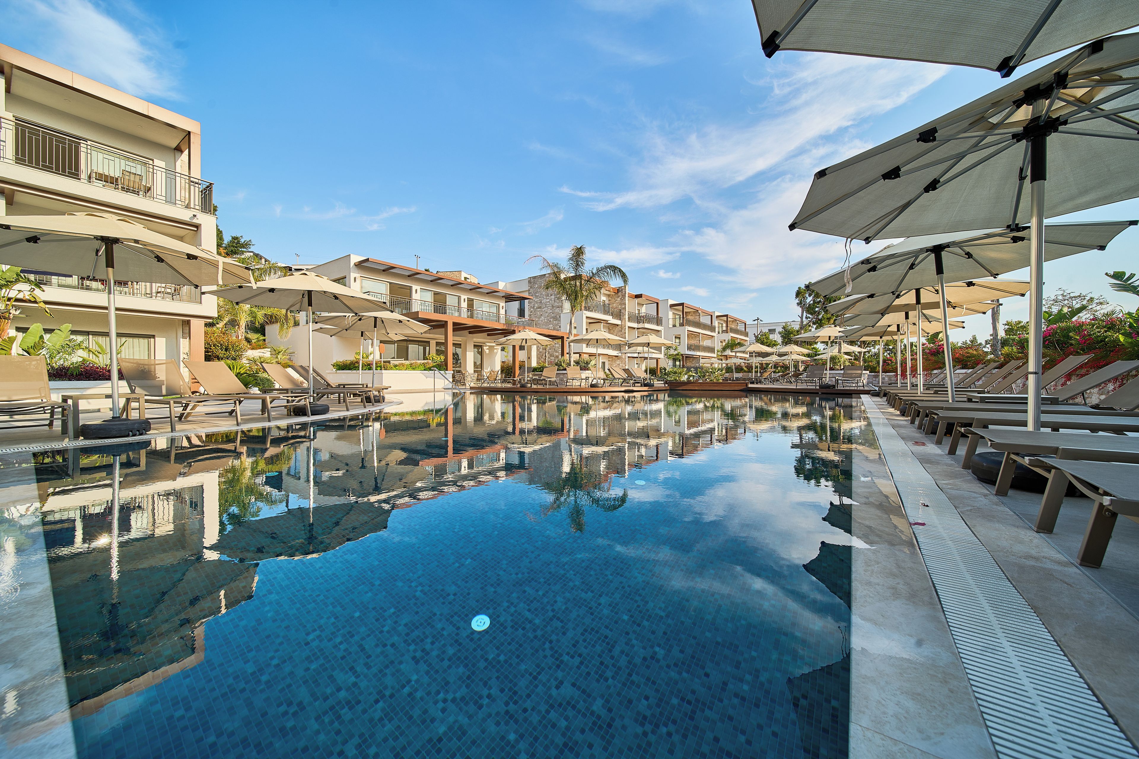 A swimming pool with a clear blue water surface, surrounded by lounge chairs and umbrellas, and adjacent to a multi-story building with balconies.