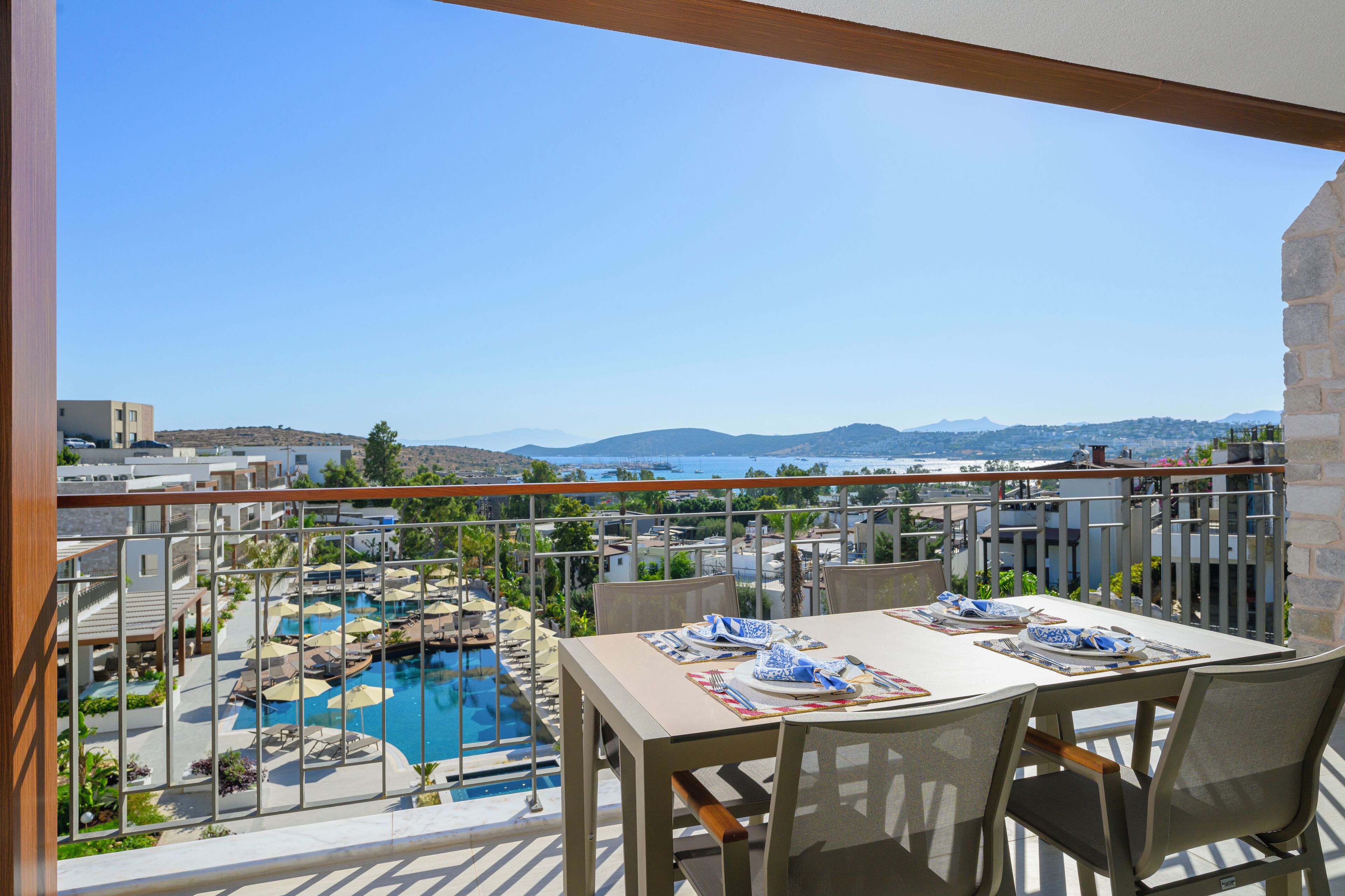 En balkon med et spisebord, udsigt over en swimmingpool og et smukt landskab med hav og bakker.