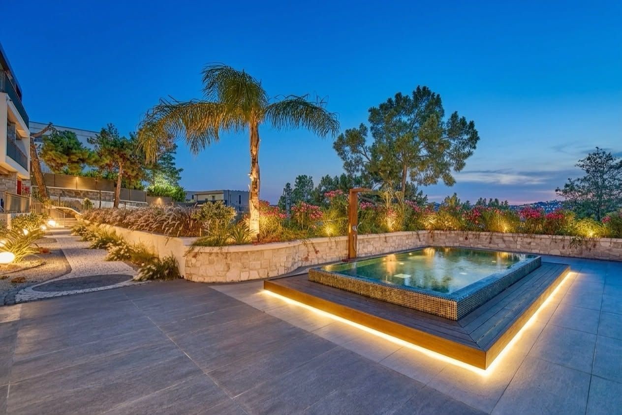 Una piscina con borde de piedra y cubierta de madera, rodeada de jardines ajardinados y palmeras.