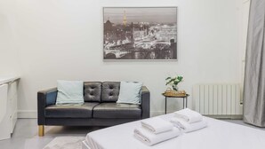 Una habitación de hotel moderna con cama, un sofá de cuero gris, un cuadro enmarcado con una vista urbana, una mesita con una planta y un radiador.